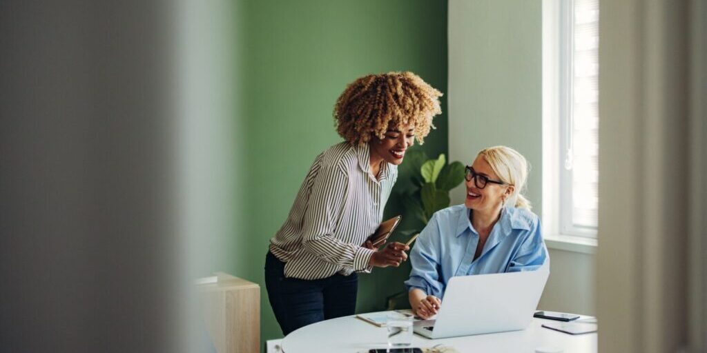 Cheerful smiling African American woman talking to female colleague who is sitting at office desk.
